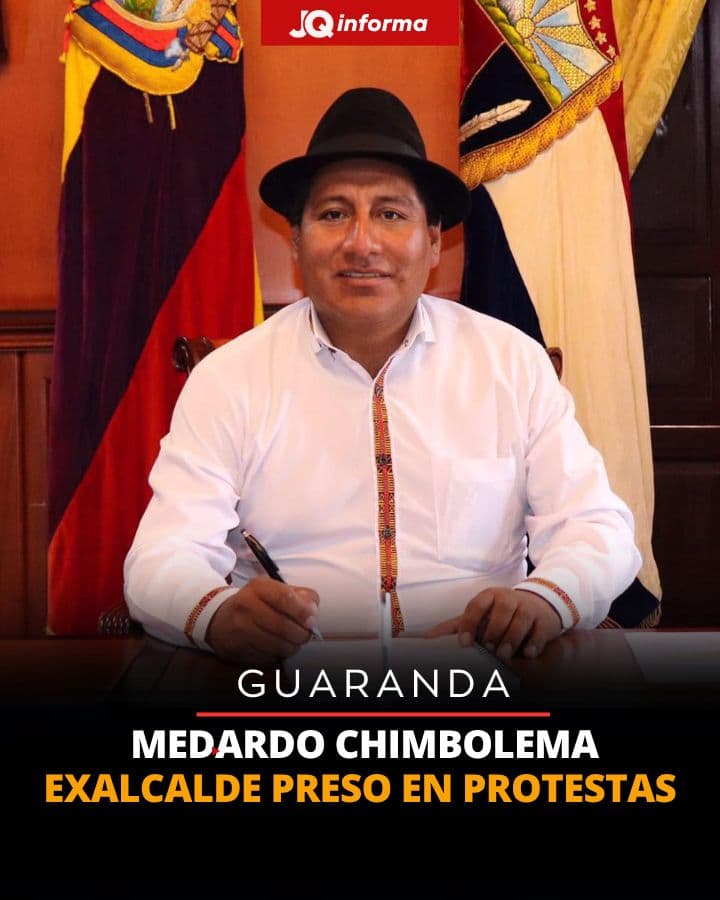 Medardo Chimbolema con sombrero negro y camisa blanca bordada, sentado frente a banderas nacionales.