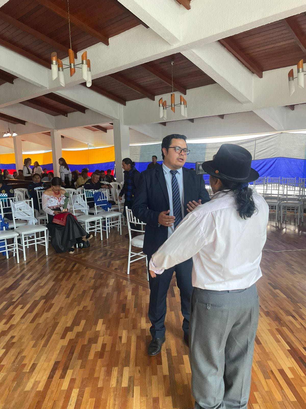 Hombre en traje formal dialoga con otro hombre con sombrero negro en un salón.