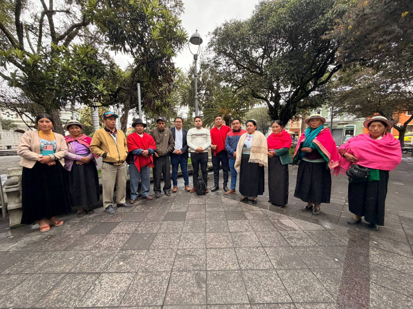 Personas con trajes tradicionales andinos y ropa moderna posando juntas en una plaza arbolada.